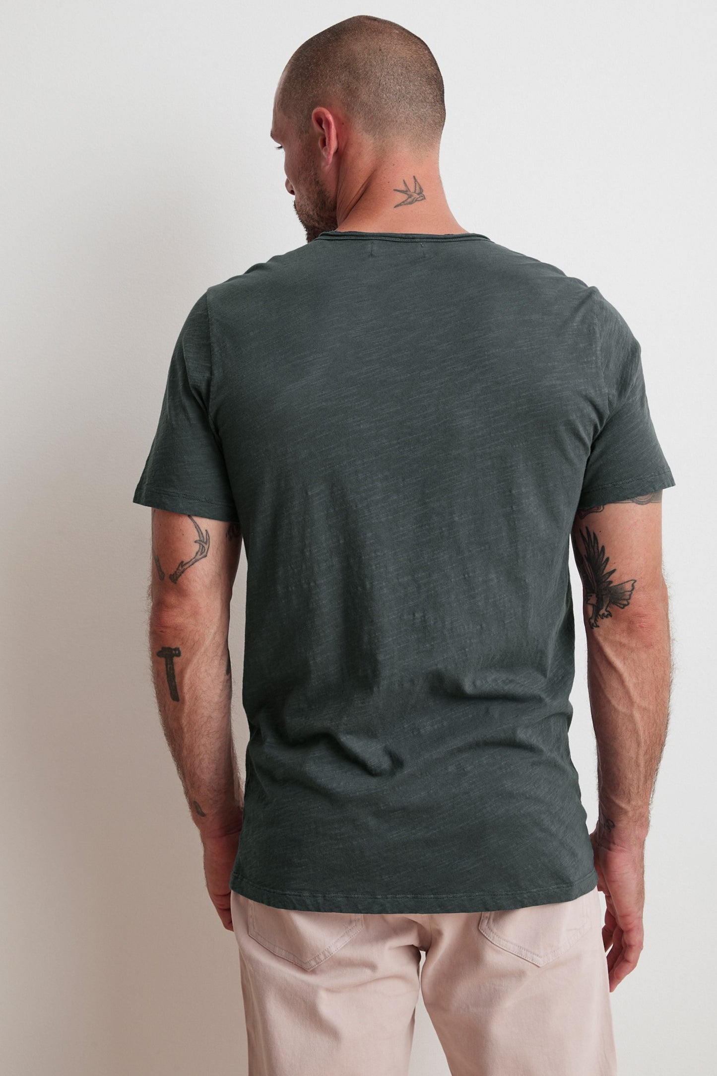 A man with tattoos on his arms is standing with his back to the camera, wearing a dark green, textured Velvet by Graham & Spencer CHAD TEE and light-colored pants.-8004715774033