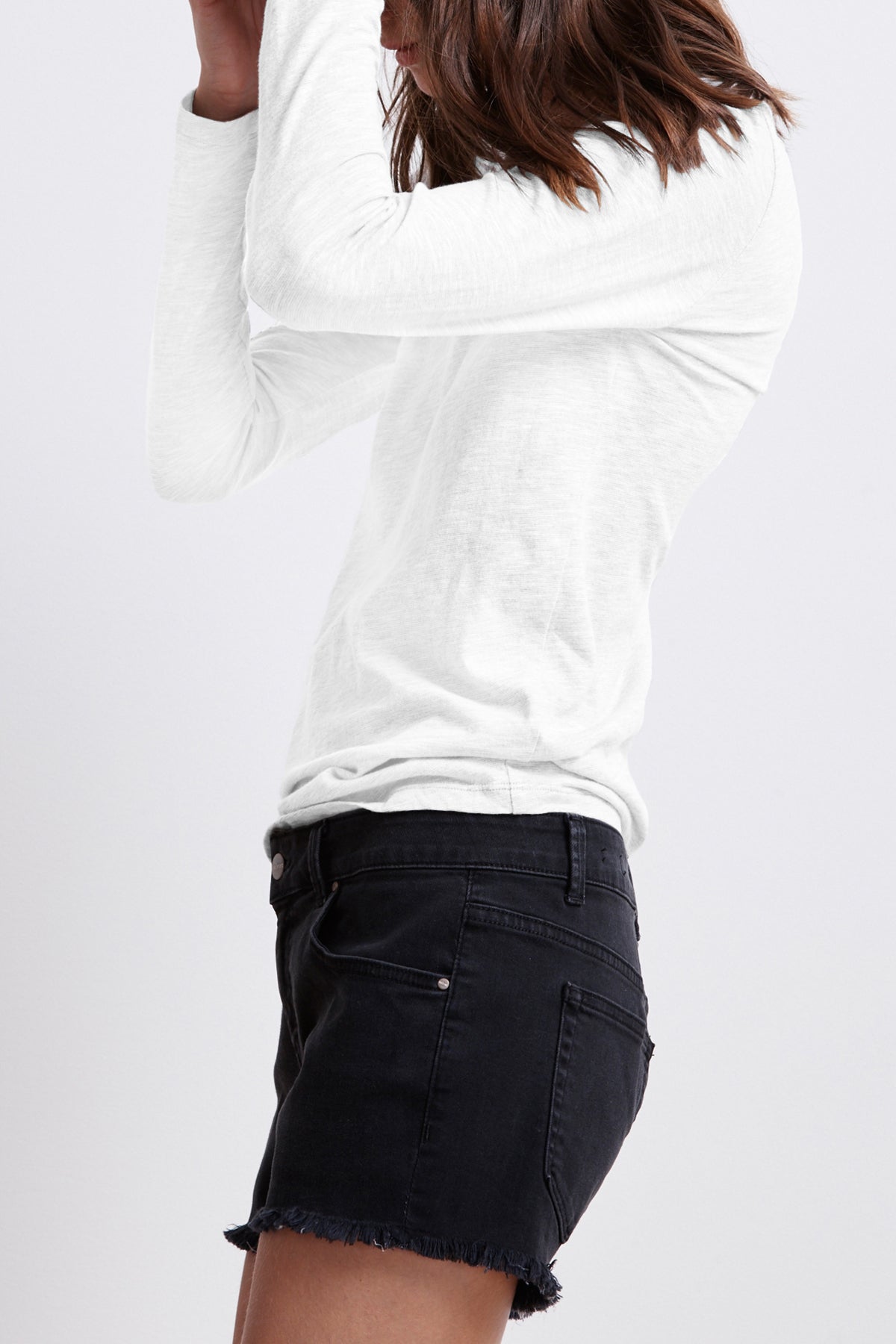 A person with shoulder-length hair, wearing a white long-sleeve shirt and the CINDY FRAYED HEM BOYFRIEND SHORTS by Velvet by Graham & Spencer, is standing and facing to the side.