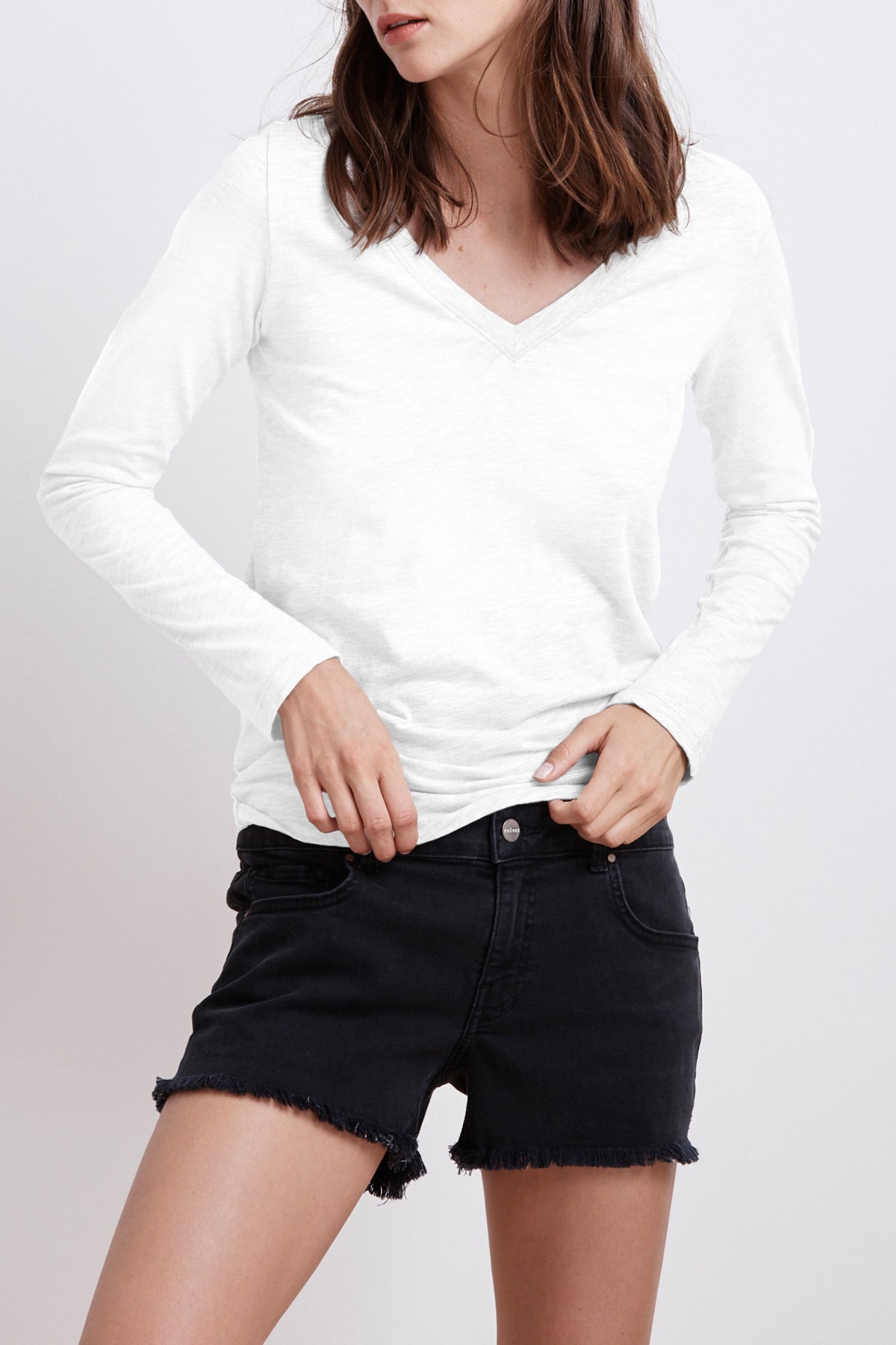 A person wearing a white V-neck long-sleeve shirt and Velvet by Graham & Spencer's CINDY FRAYED HEM BOYFRIEND SHORTS stands against a plain background. They have shoulder-length brown hair.