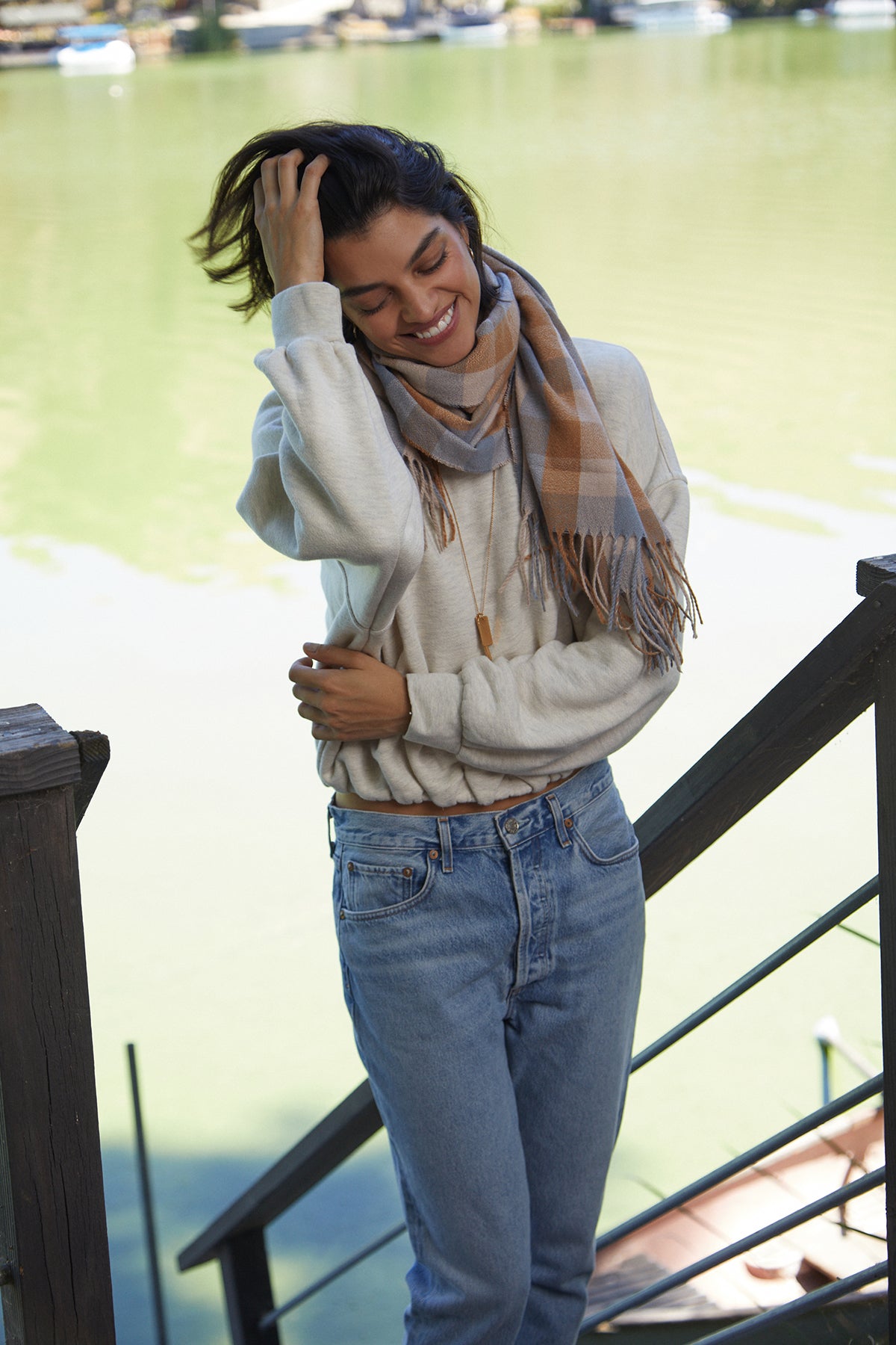 A person wearing a light sweater, jeans, and the Velvet by Graham & Spencer CHECK SCARF stands on a wooden staircase near a body of water, smiling and touching their hair.-25426487017665