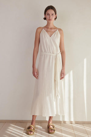 A woman in a white sleeveless CARRILLO DRESS by Velvet by Jenny Graham and gold sandals stands in a sunlit room with a neutral background.