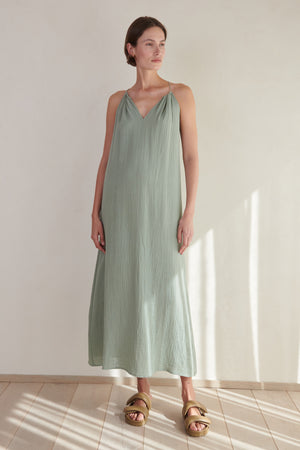 A woman stands in a sunlit room wearing a Velvet by Jenny Graham CARRILLO DRESS sleeveless dress and gold sandals.