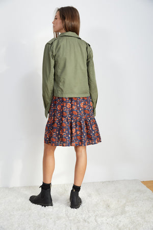 A person stands on a white carpet facing away from the camera, stylishly sporting the DIXIE CROPPED ARMY JACKET by Velvet by Graham & Spencer with a floral dress and black ankle boots, effortlessly showcasing a laid-back style.