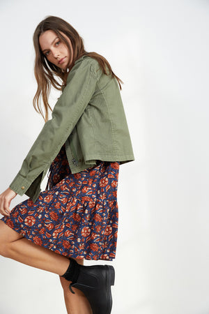 A woman with long hair in a DIXIE CROPPED ARMY JACKET by Velvet by Graham & Spencer, over a floral dress and black boots, stands against a plain white background.