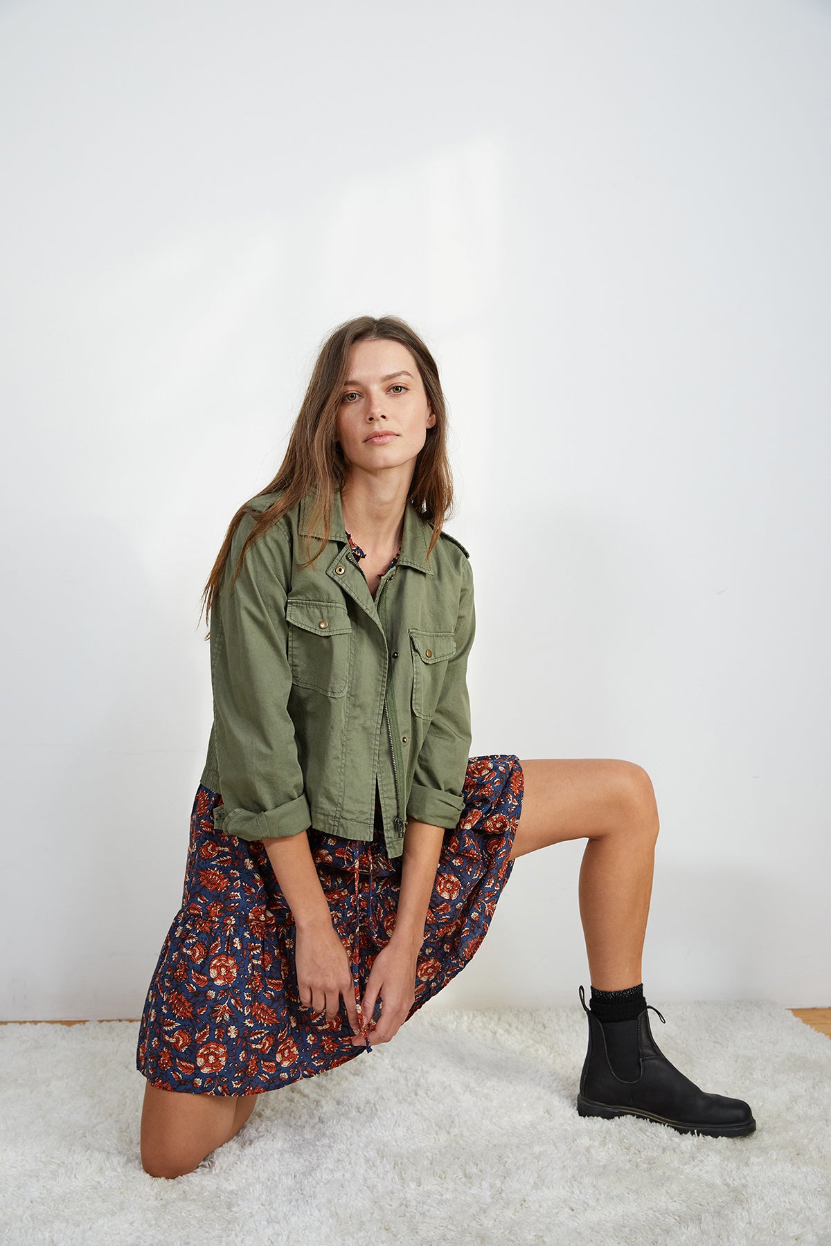 Woman kneeling on one knee, wearing a DIXIE CROPPED ARMY JACKET by Velvet by Graham & Spencer in a laid-back style, a colorful patterned skirt, and black boots, posing indoors against a plain background.-7835031699537