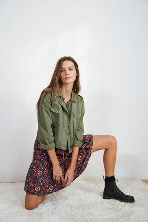 Woman kneeling on one knee, wearing a DIXIE CROPPED ARMY JACKET by Velvet by Graham & Spencer in a laid-back style, a colorful patterned skirt, and black boots, posing indoors against a plain background.
