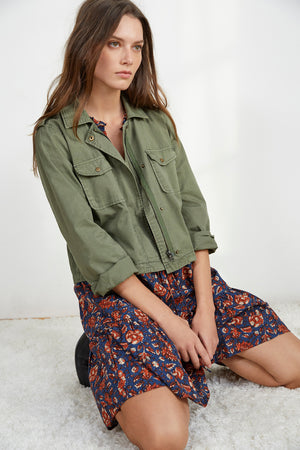 A woman with long brown hair sits on the floor, wearing the DIXIE CROPPED ARMY JACKET from Velvet by Graham & Spencer over a floral dress, her laid-back style evident as she gazes off to the side.
