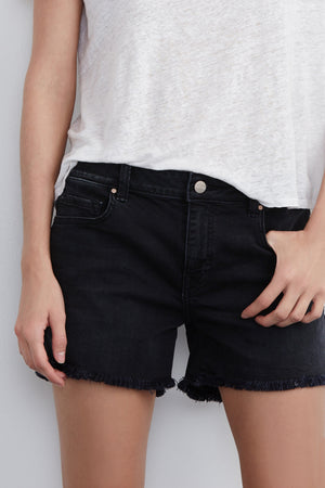 A person is wearing a white t-shirt and Velvet by Graham & Spencer's CINDY FRAYED HEM BOYFRIEND SHORTS in black, standing against a plain background. They have one hand in their pocket, exuding a relaxed silhouette.