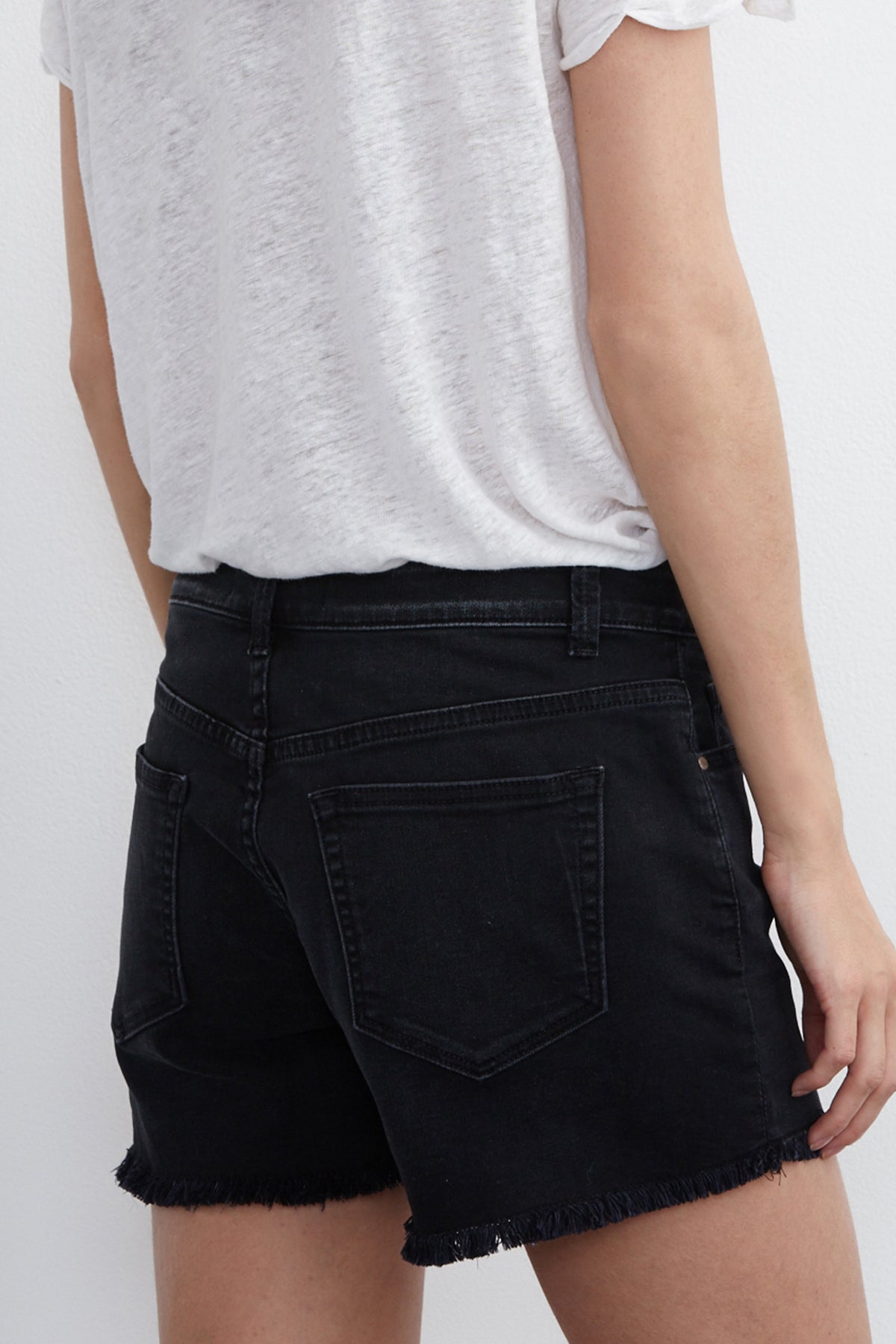 A person wearing a white t-shirt and Velvet by Graham & Spencer’s CINDY FRAYED HEM BOYFRIEND SHORTS in black, photographed from the back, showcases a relaxed silhouette.-17336149704897