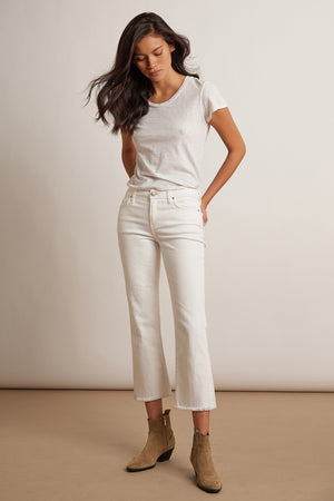 A woman stands against a plain background wearing a white t-shirt, Velvet by Graham & Spencer TWIGGY HIGH RISE CROP JEAN in white, and tan ankle boots.