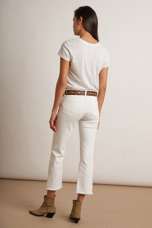A woman stands facing away, wearing a white t-shirt, the TWIGGY HIGH RISE CROP JEAN by Velvet by Graham & Spencer featuring a frayed hem, a brown belt, and tan ankle boots.