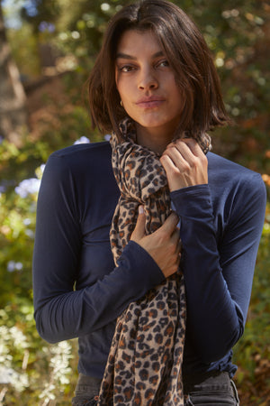 A person with short dark hair, wearing a long-sleeve navy top and a MINI LEOPARD SCARF by Velvet by Graham & Spencer, stands outdoors in a natural setting.