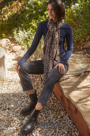 A person sits outdoors on a stone ledge, wearing a long-sleeve navy shirt, dark jeans, black boots, and a MINI LEOPARD SCARF by Velvet by Graham & Spencer. The background features greenery and a path of small stones.