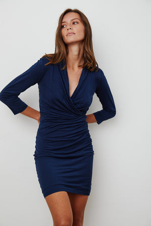 A person poses in a fitted, ROZA GAUZY WHISPER 3/4 SLEEVE WRAP DRESS by Velvet by Graham & Spencer, an ideal figure flatterer against a plain background.
