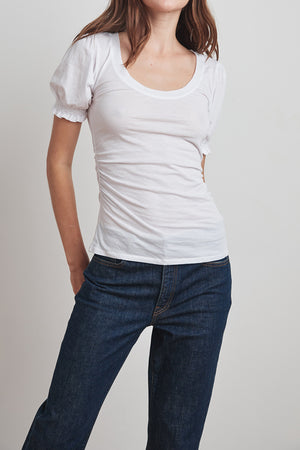 A person wearing a white Velvet by Graham & Spencer KAMI GAUZY WHISPER SHIRRED SLEEVE TOP and dark blue jeans is standing against a plain white background.