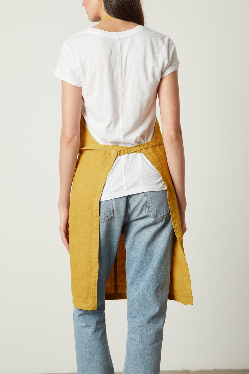 A woman from the back wearing a white t-shirt and blue jeans with a Jenny Graham Home linen apron tied around her waist.