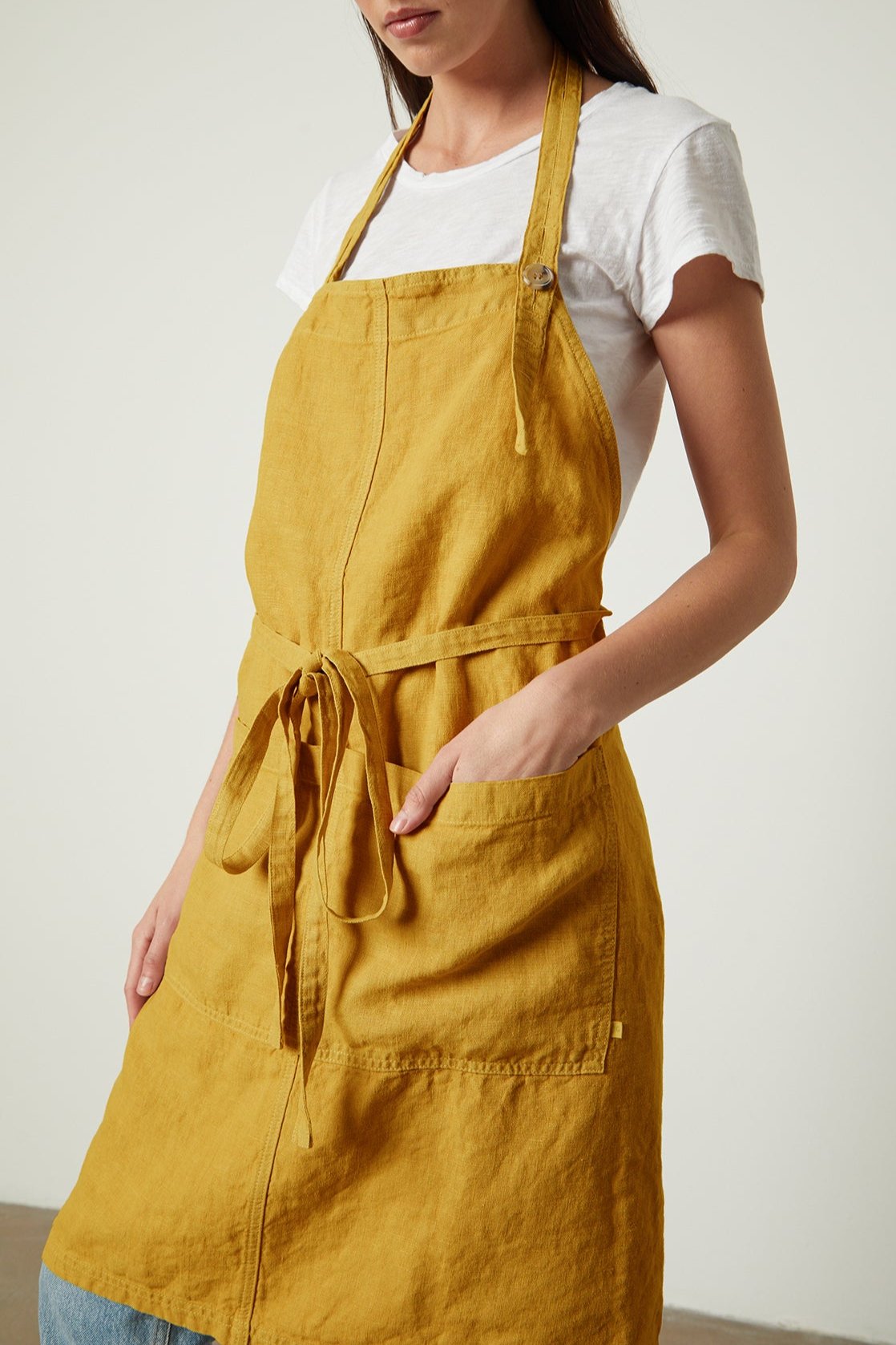 A woman in a white t-shirt and a durable Jenny Graham Home linen apron stands thoughtfully, her head tilted down and to the side.-25519630090433