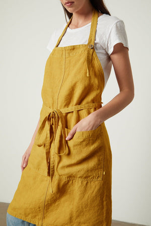 A woman in a white t-shirt and a durable Jenny Graham Home linen apron stands thoughtfully, her head tilted down and to the side.