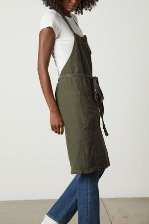 Person wearing a white t-shirt, Jenny Graham Home LINEN APRON with an adjustable neck strap, and blue jeans, standing against a plain background. They have curly hair and are looking down to their right.