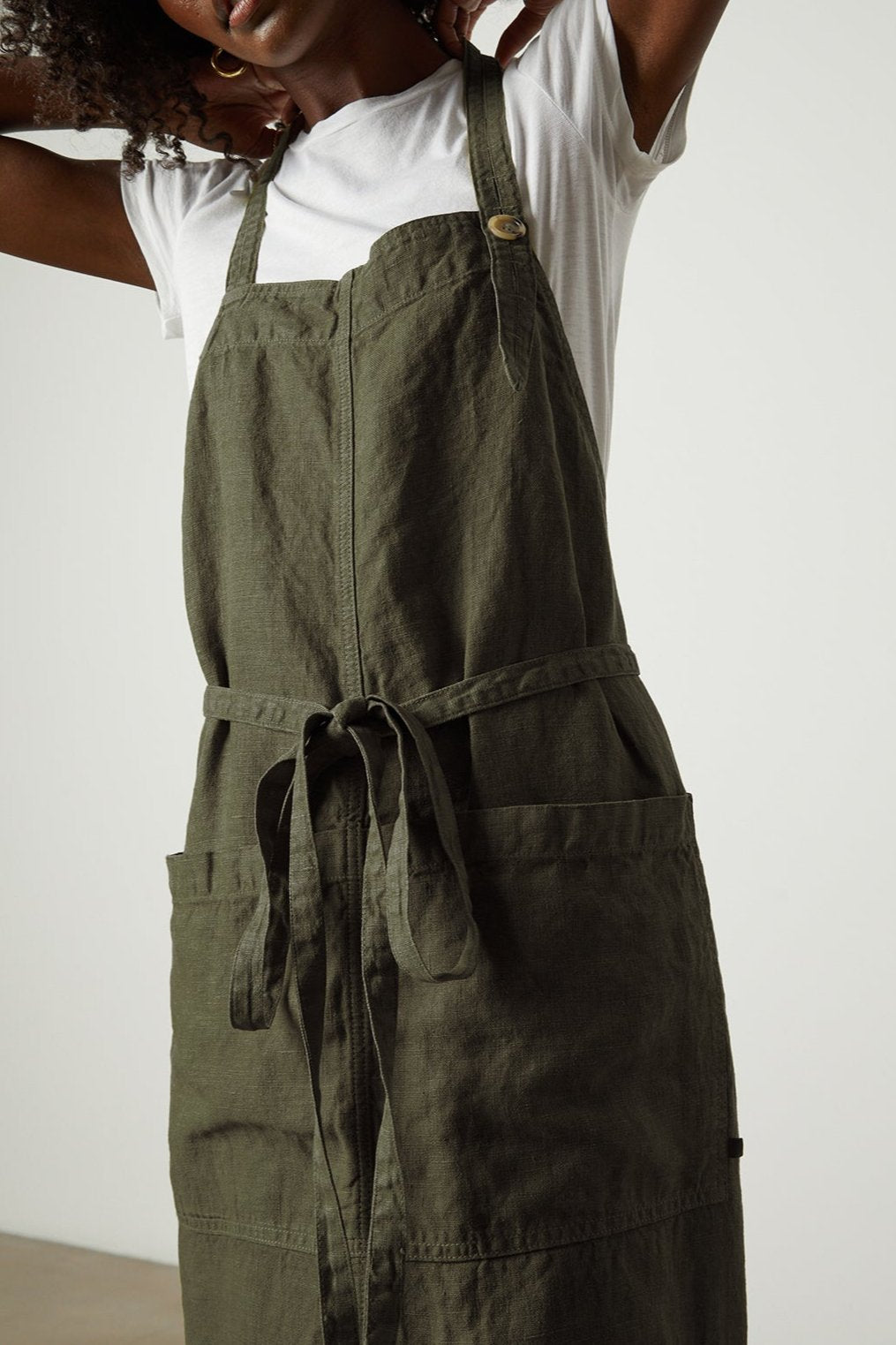 Person wearing a white t-shirt and a green Jenny Graham Home LINEN APRON with large pockets adjusts the adjustable neck strap.-25519629205697