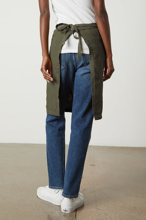 Person standing with their back to the camera, wearing a white t-shirt, jeans, white sneakers, and a dark green unisex LINEN APRON by Jenny Graham Home with an adjustable neck strap tied around the waist.