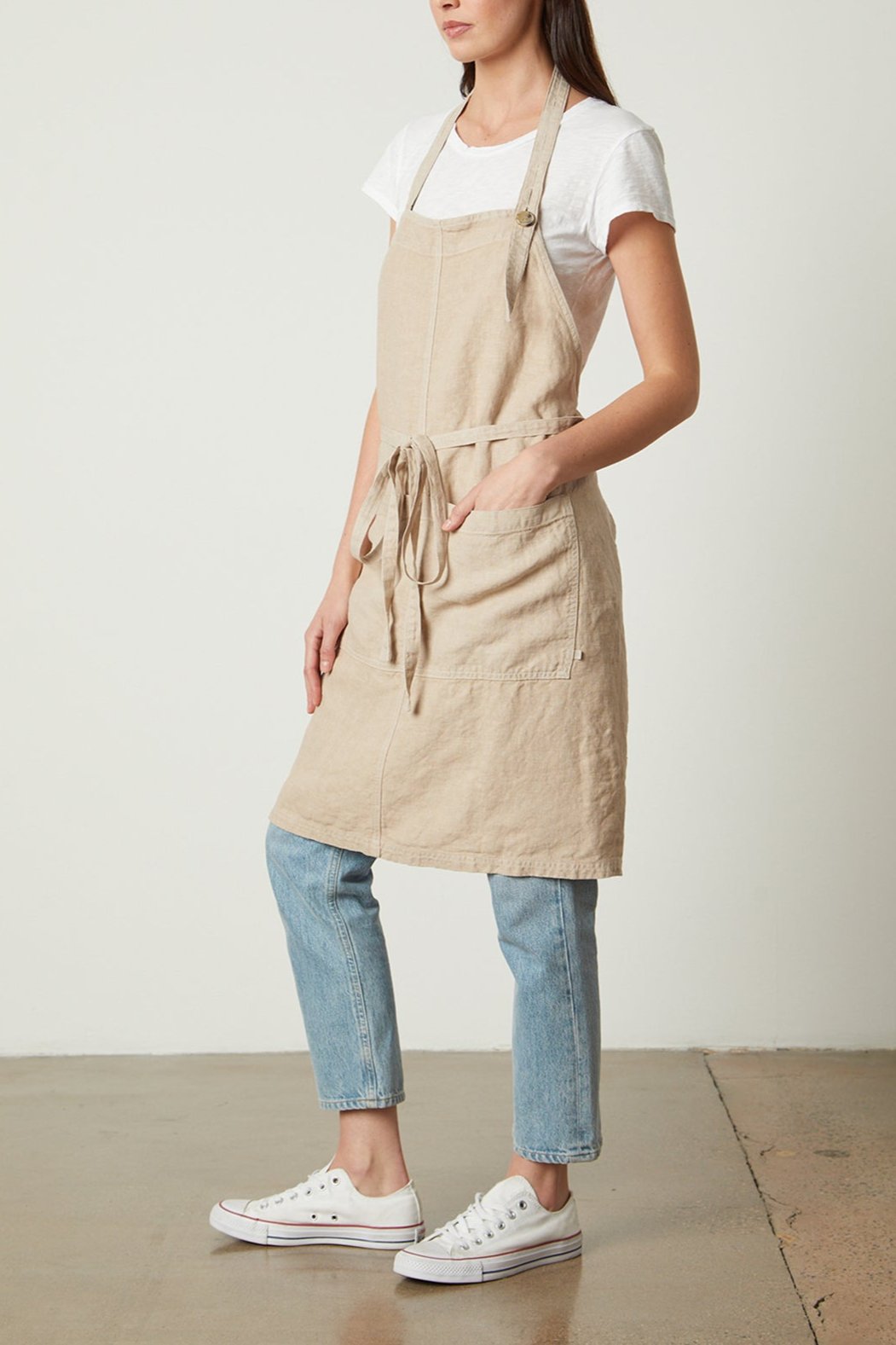 A woman stands against a plain background wearing a beige, unisex Jenny Graham Home LINEN APRON with an adjustable neck strap over a white t-shirt and blue jeans. She also wears white sneakers and has her hands in the durable linen apron pockets.-25519629041857