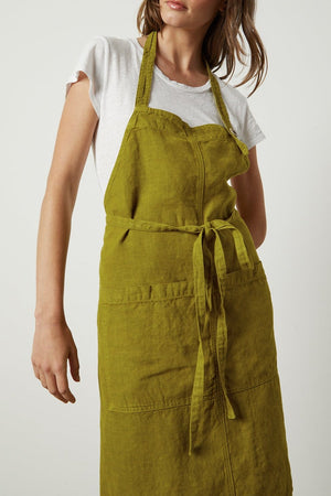 A woman in a white t-shirt and Jenny Graham Home LINEN APRON with an adjustable neck strap stands against a neutral background, looking down to her right with a relaxed expression.
