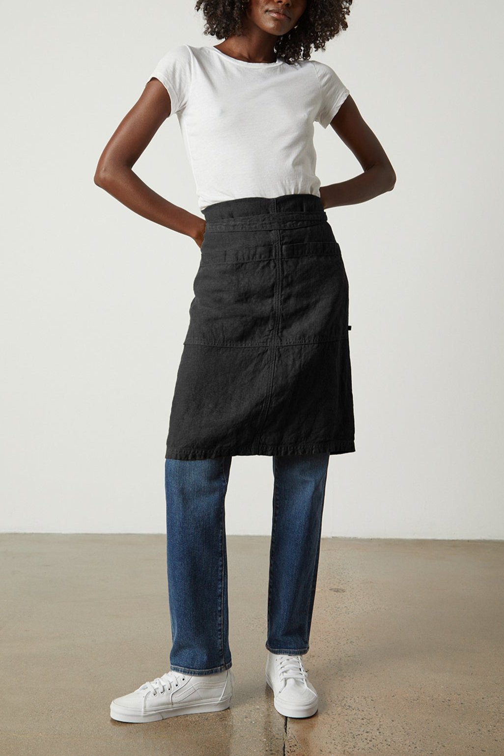 A person with curly hair stands against a plain background, wearing a white t-shirt, blue jeans, a Jenny Graham Home LINEN APRON with an adjustable neck strap, and white sneakers.-25519629664449