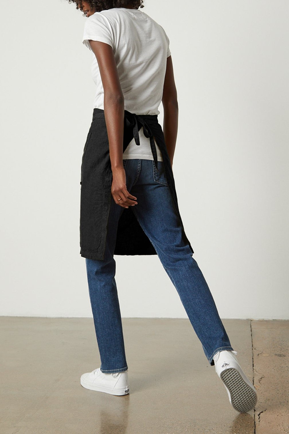 A person with curly hair, wearing a white T-shirt, blue jeans, white sneakers, and a black LINEN APRON from Jenny Graham Home with an adjustable neck strap, stands with their back to the camera in an indoor setting.-25519629795521