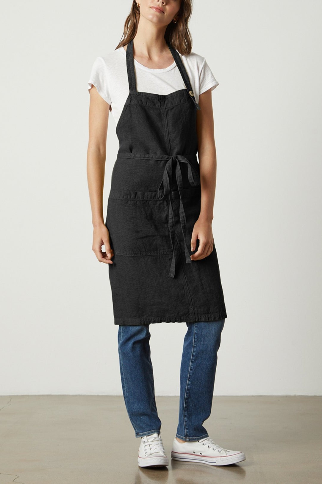 A person stands against a plain background wearing the LINEN APRON by Jenny Graham Home, a durable linen, unisex apron with an adjustable neck strap over a white t-shirt and blue jeans, paired with white sneakers.