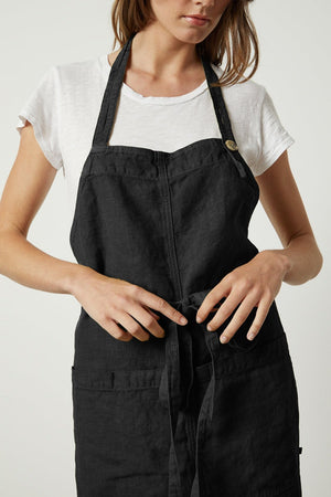 A person wearing a white T-shirt and a black unisex apron with an adjustable neck strap is tying the waist straps of the durable LINEN APRON by Jenny Graham Home.