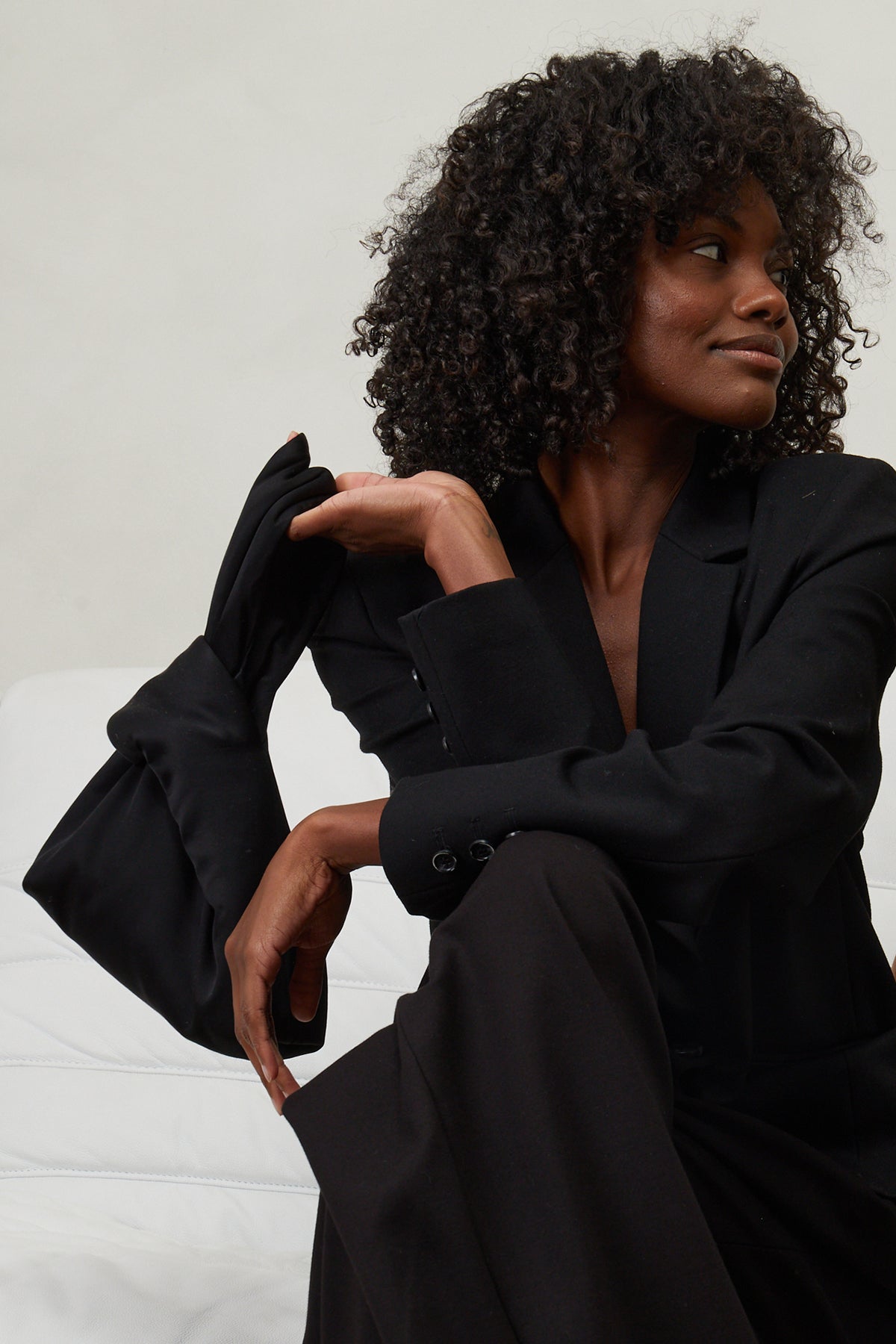 A woman in a black blazer and pants sitting on a bed, holding the Jenny Bag by Velvet by Jenny Graham.