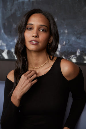 A person with long dark hair wears a black LINNIE CUT-OUT DRESS by Velvet by Graham & Spencer, accessorized with a gold necklace and hoop earrings, posing in front of a dark, abstract art background.