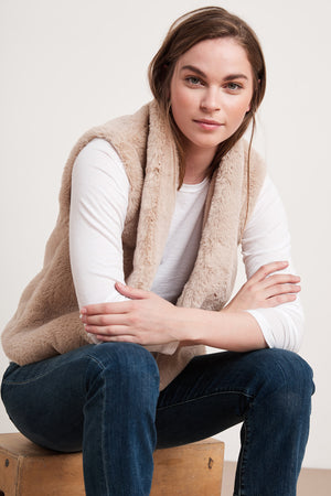 A woman seated on a wooden box wears a white long-sleeve shirt paired with the CHELLE FAUX LUX FUR OPEN VEST by Velvet by Graham & Spencer, her arms crossed and her expression neutral. This versatile layering piece is perfect for the cooler months.