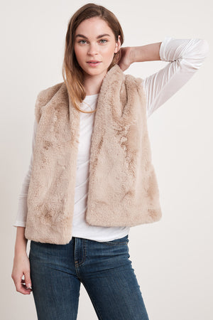 A woman in a white long-sleeve shirt and blue jeans poses while wearing the CHELLE FAUX LUX FUR OPEN VEST by Velvet by Graham & Spencer, an eco-chic piece perfect for layering during the cooler months.