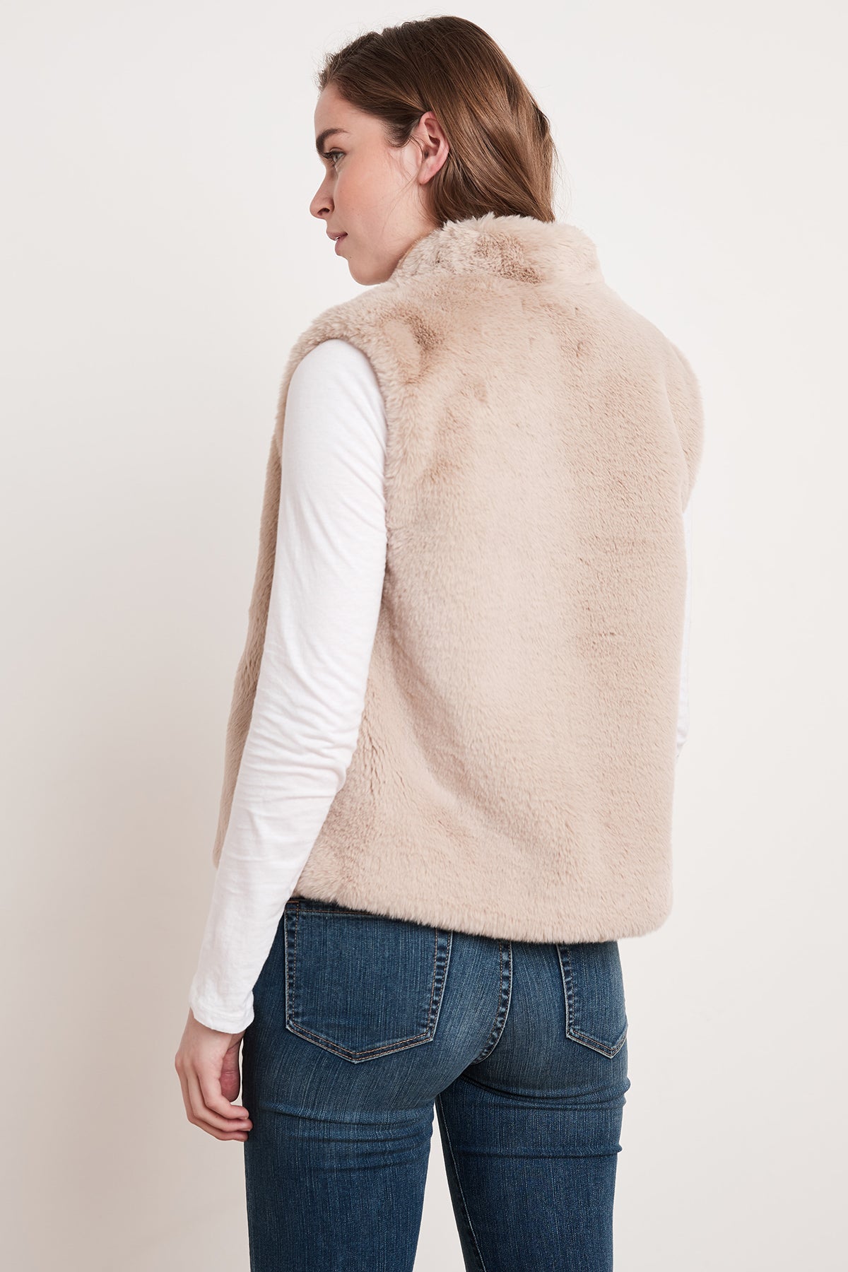 A woman wearing the CHELLE FAUX LUX FUR OPEN VEST from Velvet by Graham & Spencer, layered over a white long-sleeve shirt, stands with her back turned, looking to the side. This stylish vest is perfect for the cooler months.-5697530232913