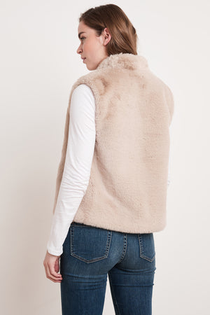 A woman wearing the CHELLE FAUX LUX FUR OPEN VEST from Velvet by Graham & Spencer, layered over a white long-sleeve shirt, stands with her back turned, looking to the side. This stylish vest is perfect for the cooler months.