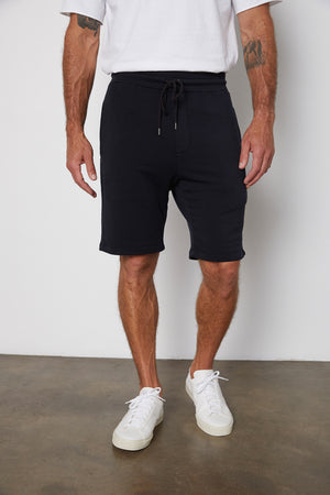 A man wearing a white T-shirt, Velvet by Graham & Spencer's ATLAS SHORT with a tailored design, and white sneakers stands on a plain floor with his hands by his sides.