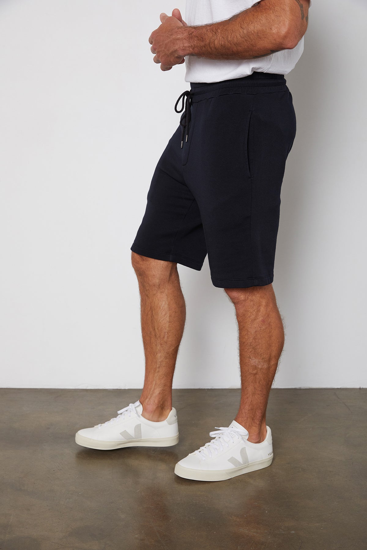 A person wearing a white t-shirt, navy Velvet by Graham & Spencer ATLAS SHORT, and white sneakers is standing in a room with a concrete floor. The image is cropped to show from the shoulders down.-25578322985153