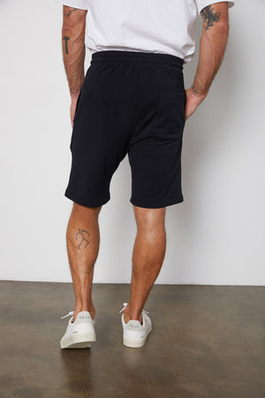 A person is standing with their back to the camera, wearing a white shirt, black Velvet by Graham & Spencer ATLAS SHORT, and white Veja sneakers. They have tattoos on their arms and one on their lower right leg.