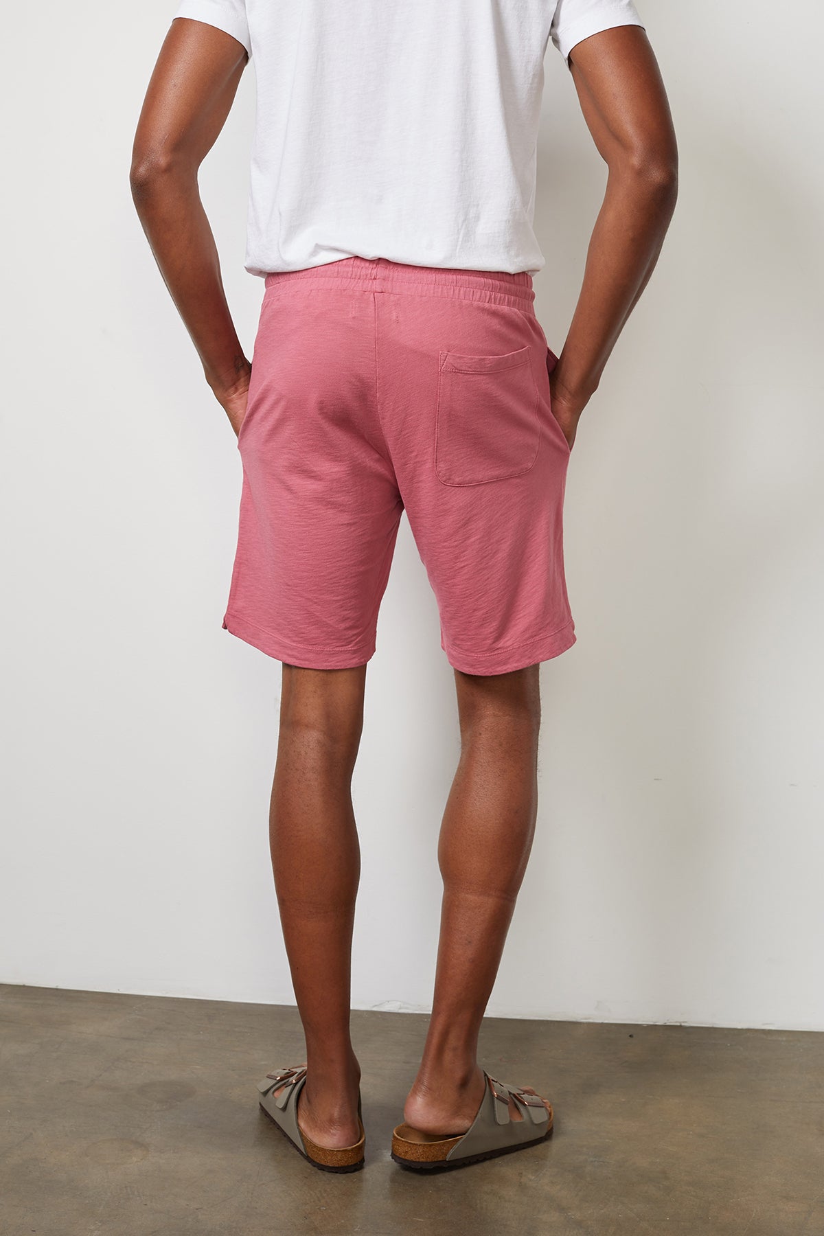 A person wearing a white t-shirt, Velvet by Graham & Spencer's CYRUS MICRO TERRY SHORT with an elastic waist, and sandals stands facing a wall with hands in pockets.-15059521503425