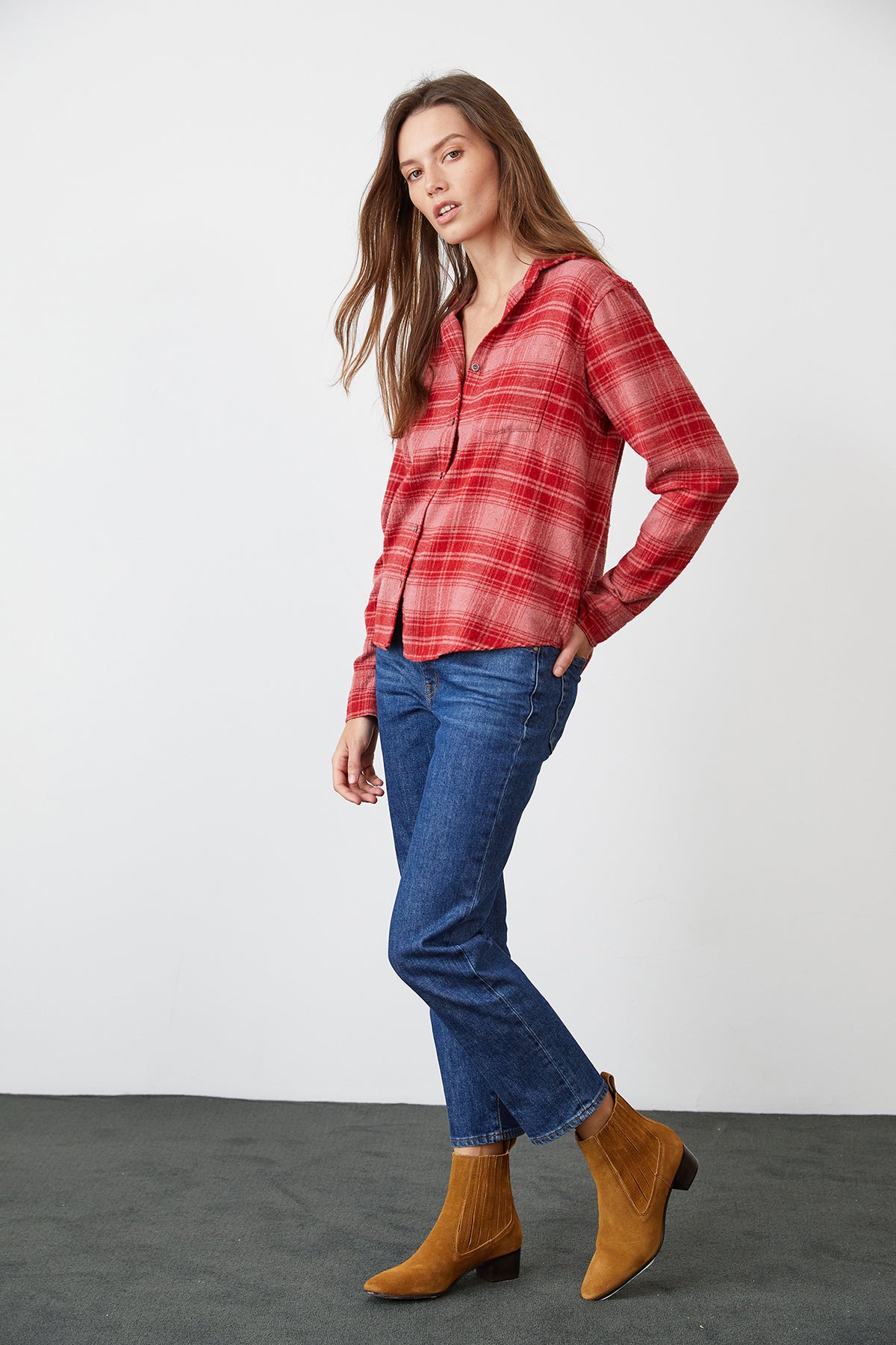 A person with long hair wearing a Velvet by Graham & Spencer RUZ Plaid Button-Up Shirt in red, classic button-up front, blue jeans, and brown boots stands against a plain background with one hand in their pocket.