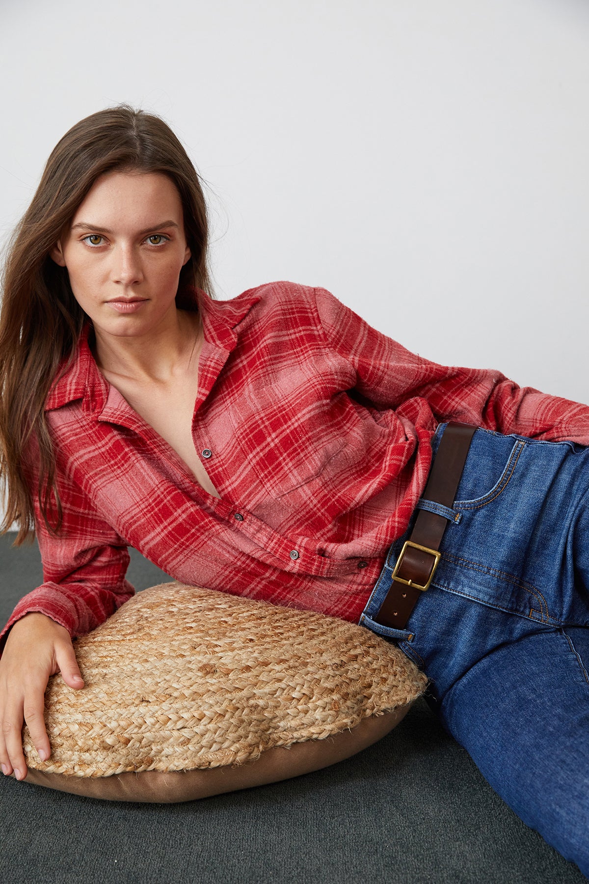 A person with long hair wearing a Velvet by Graham & Spencer RUZ PLAID BUTTON-UP SHIRT and blue jeans is reclining on a woven cushion, against a plain white background.