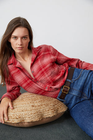 A person with long hair wearing a Velvet by Graham & Spencer RUZ PLAID BUTTON-UP SHIRT and blue jeans is reclining on a woven cushion, against a plain white background.