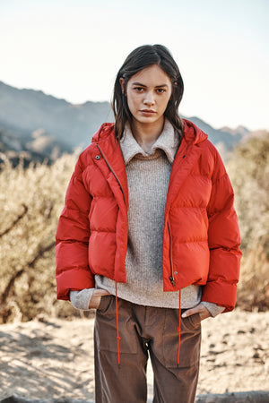 A person stands outdoors in a cozy red RAYLIN CROPPED PUFFER JACKET by Velvet by Graham & Spencer over a gray sweater and brown pants, with a mountainous landscape in the background.