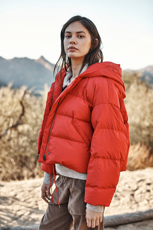 A person with long hair stands outdoors in a cozy red RAYLIN CROPPED PUFFER JACKET by Velvet by Graham & Spencer, looking at the camera. Mountains and trees provide a stunning background.