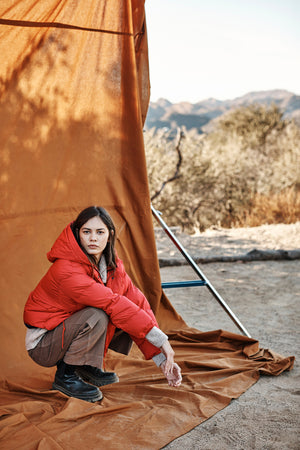 A person wearing a red Velvet by Graham & Spencer RAYLIN CROPPED PUFFER JACKET and brown pants crouches on an orange tarp outdoors, with a metal frame and desert landscape in the background.