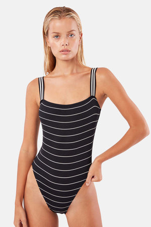 A woman models the RILEY Pinstripe Ribbed One Piece by Solid & Striped, featuring classic black-and-white stripes, against a plain background.