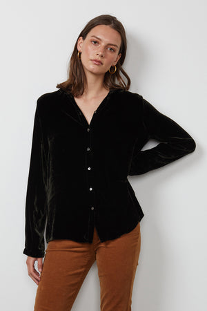 A person with shoulder-length hair wearing a JENSINE Silk Velvet Button-Up Shirt by Velvet by Graham & Spencer and brown pants stands against a plain white background.