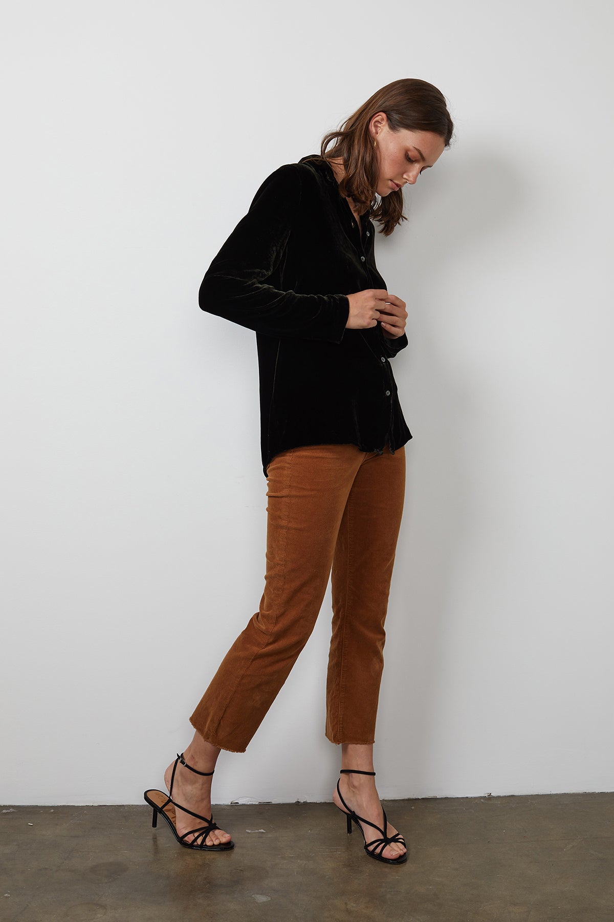 A person wearing the JENSINE SILK VELVET BUTTON-UP SHIRT in black from Velvet by Graham & Spencer, paired with brown pants and black strappy heels, adjusts their top while facing slightly to the right against a plain white background, subtly hinting at the smooth elegance of velvet in their ensemble.-15201374601409